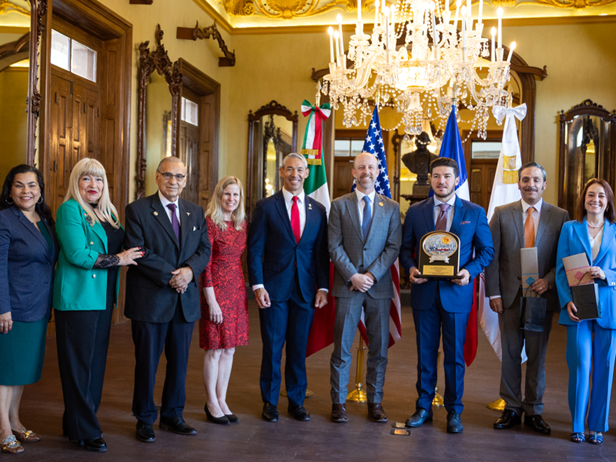 Delegacin binacional posa en el Saln Jurez tras la reunin de alcaldes del sur de Texas en Monterrey al centro representantes texanos y de Nuevo Len sostienen reconocimientos flanqueados por banderas de Mxico Estados Unidos y Nuevo Len bajo una lmpara de cristal
