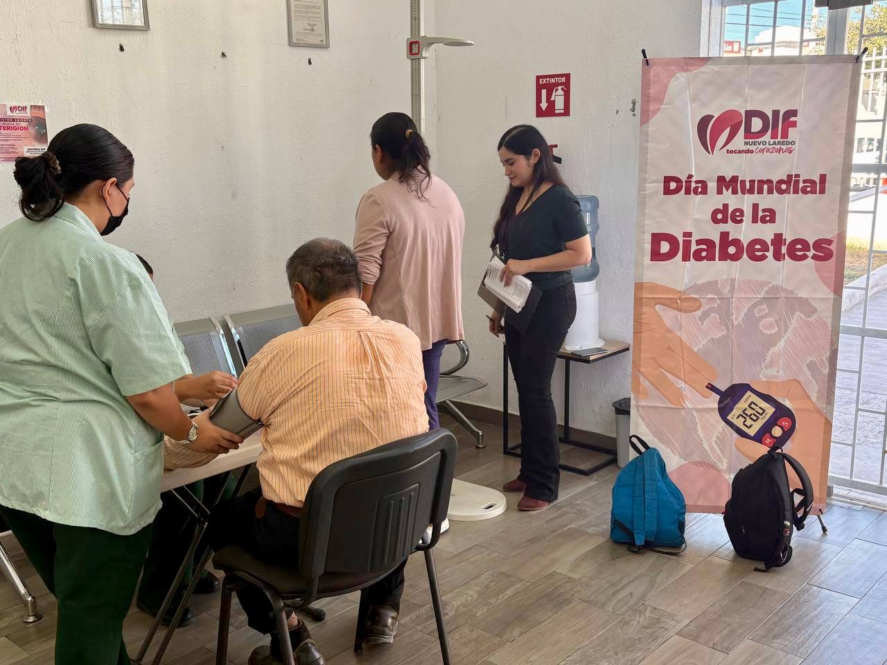 Varias personas participan en una jornada del DIF por el Da Mundial de la Diabetes una mujer toma la presin a un hombre junto a un cartel informativo del evento