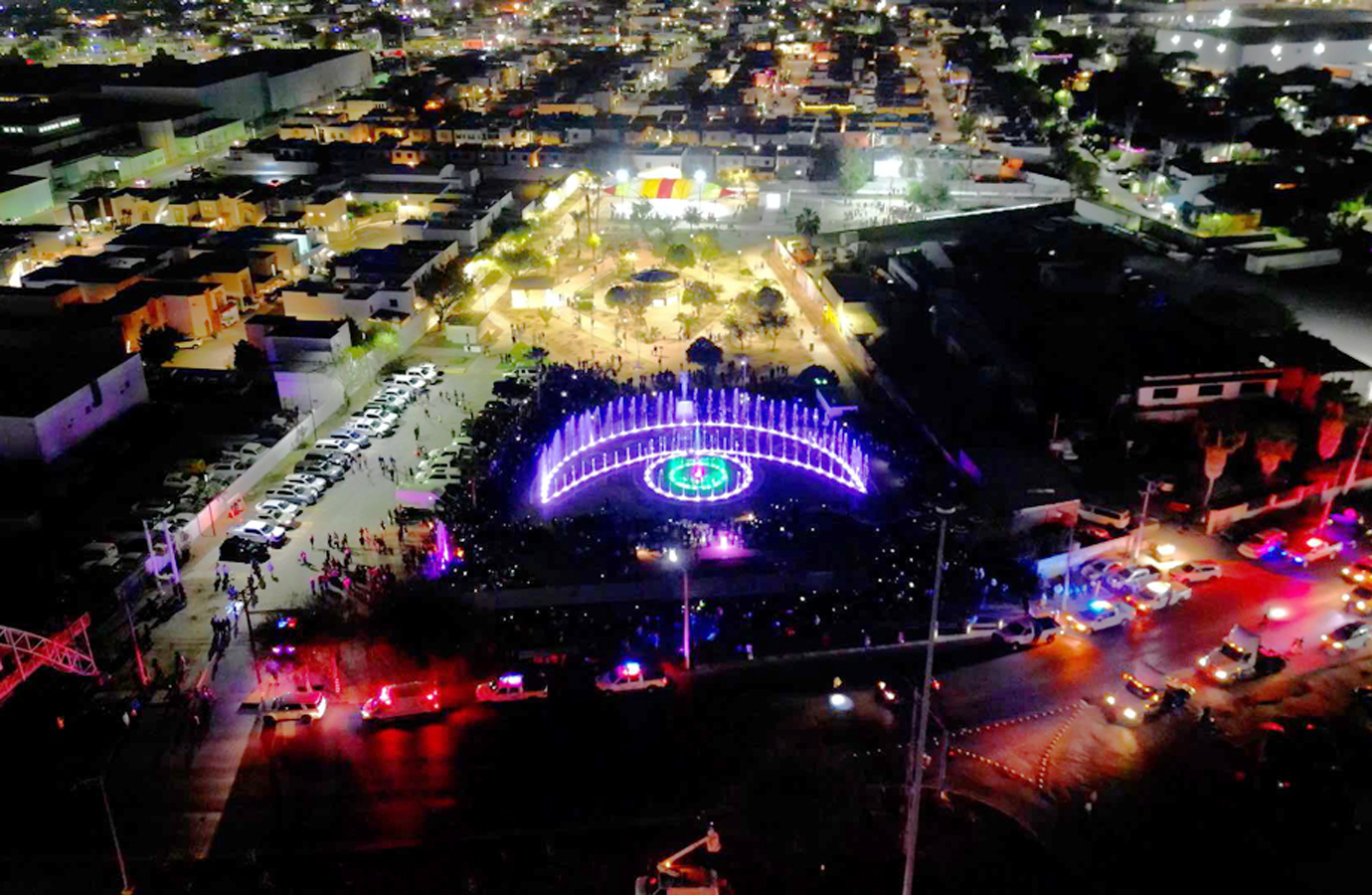 Vista aérea nocturna de la Plaza de la Mujer en Nuevo Laredo con fuente danzante iluminada y familias reunidas.
