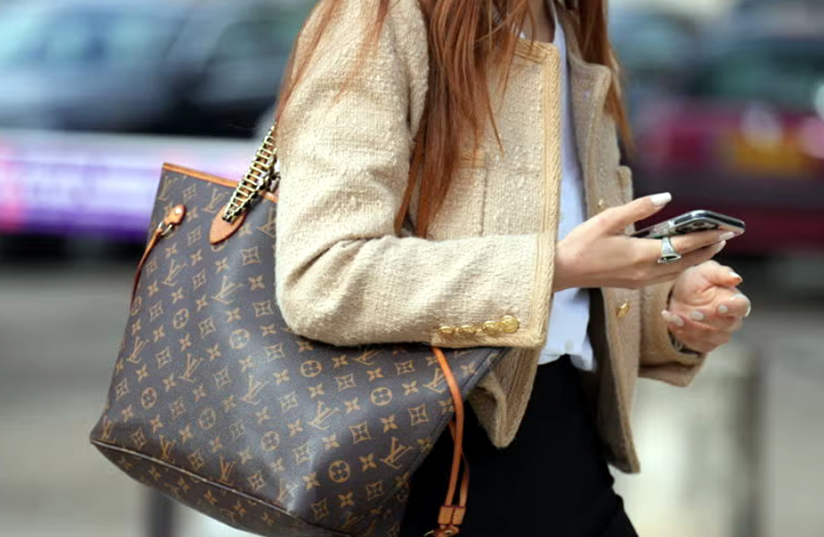 Mujer caminando con un bolso Louis Vuitton en el brazo mientras revisa su teléfono celular; imagen ilustrativa relacionada con una disputa doméstica en Laredo por bolsos extraviados.