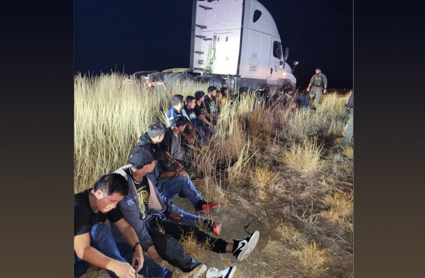 Migrantes sentados en el pastizal junto a un tráiler blanco detenido de noche en La Salle County, Texas, mientras agentes armados de DPS y la Patrulla Fronteriza aseguran la zona.