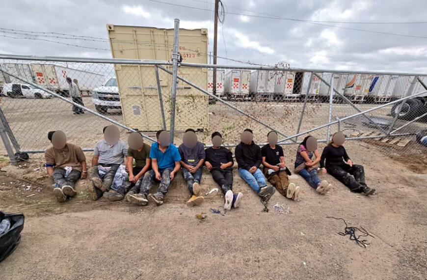 Diez personas migrantes sentadas en el suelo junto a una barda derribada en Las Minas, bajo custodia de agentes, con los rostros difuminados.