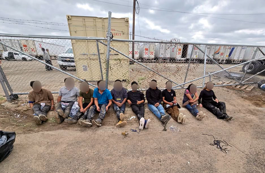 Diez personas migrantes sentadas en el suelo junto a una barda derribada en Las Minas, bajo custodia de agentes, con los rostros difuminados.