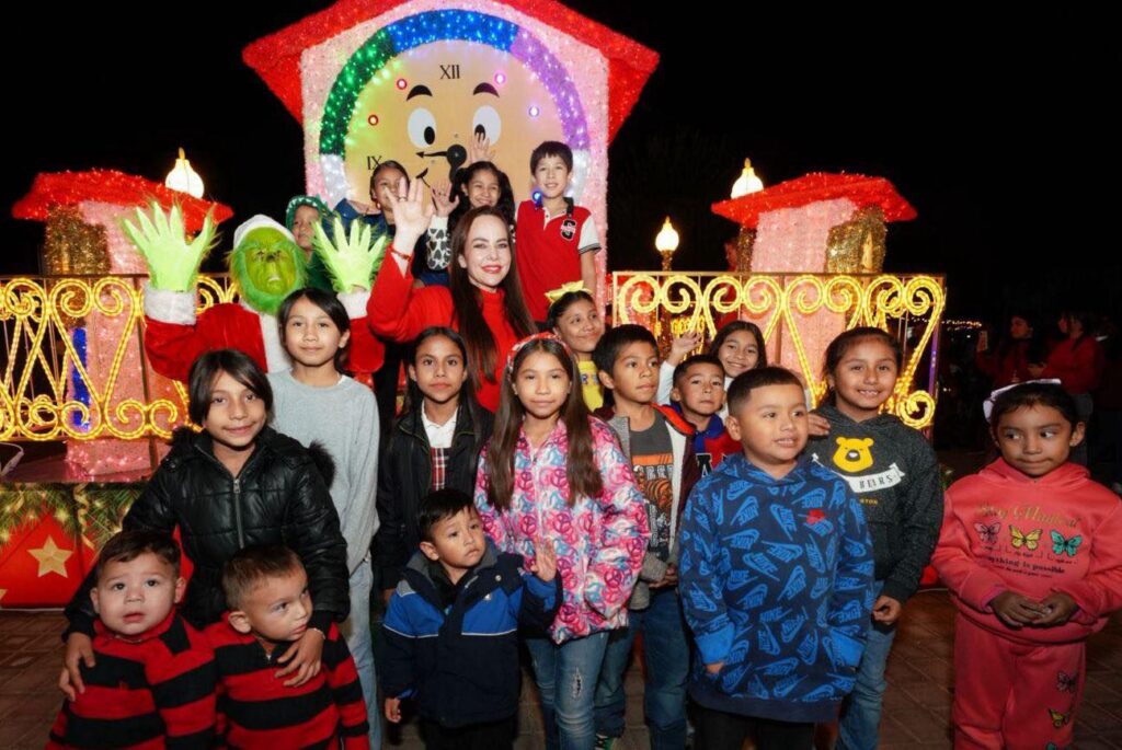 Niñas y niños junto a la alcaldesa y un Grinch disfrazado frente a un reloj navideño gigante lleno de luces.