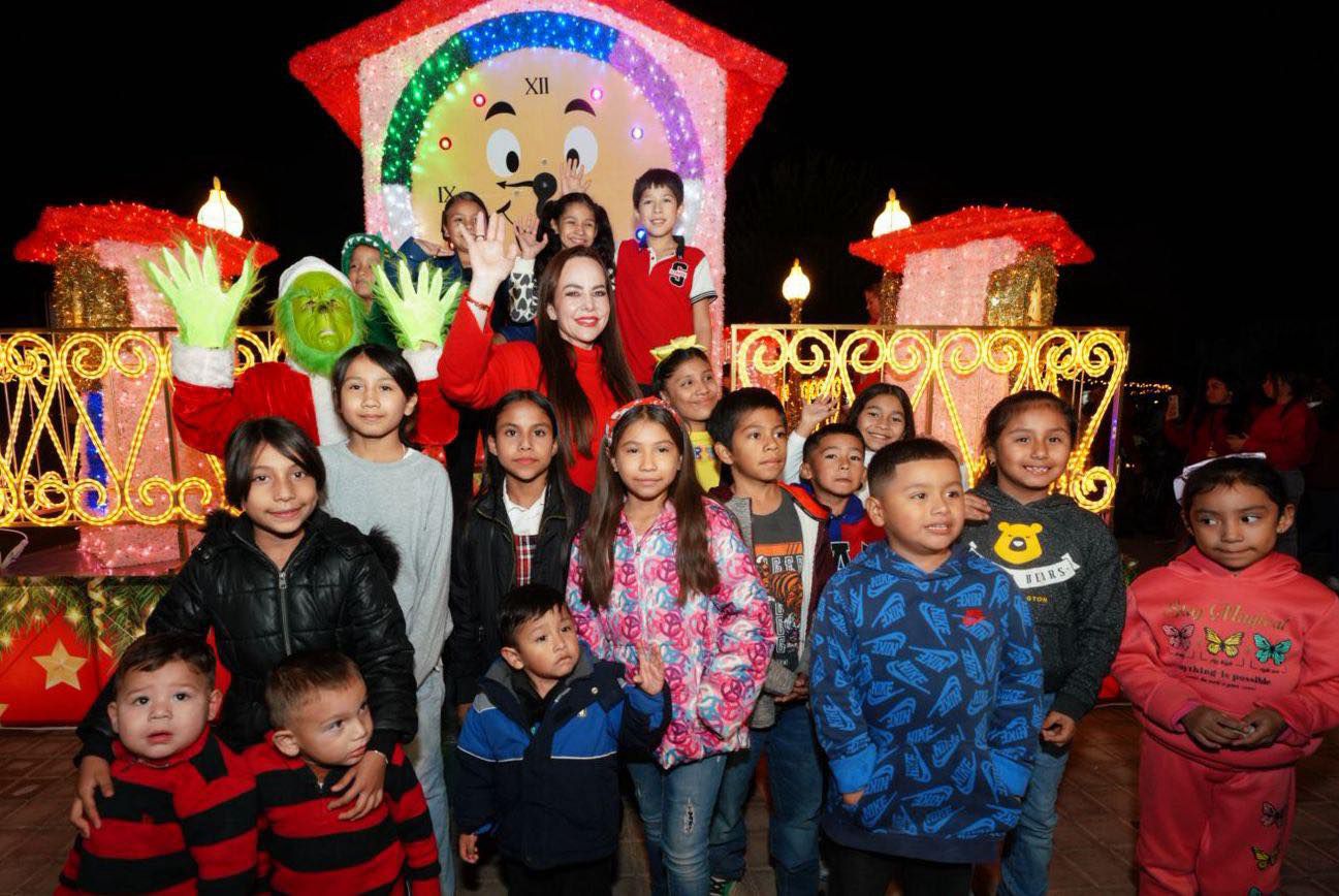 Niñas y niños junto a la alcaldesa y un Grinch disfrazado frente a un reloj navideño gigante lleno de luces.