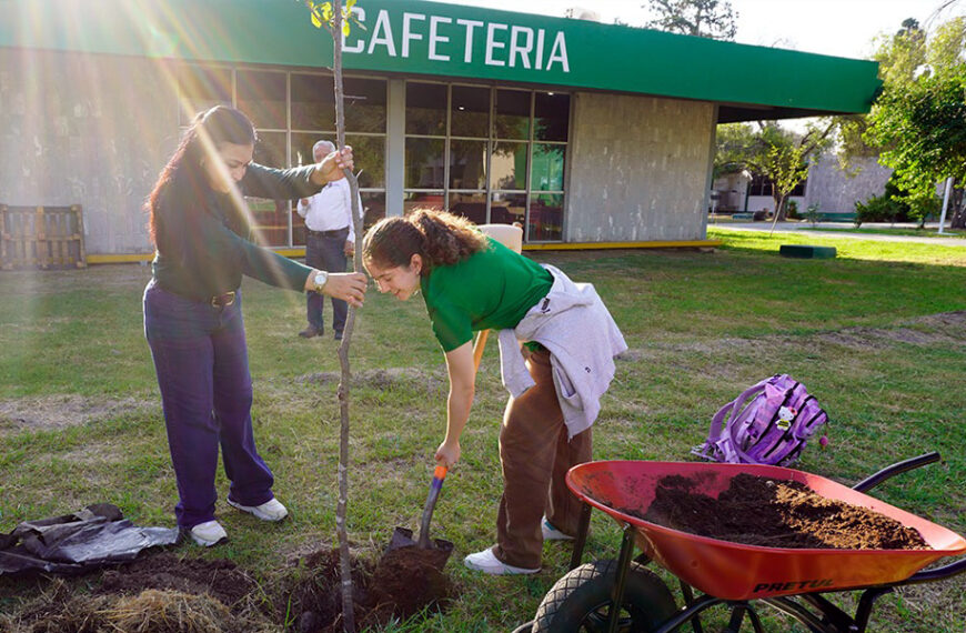 Dos personas plantan un árbol en el área verde frente al edificio de la cafetería del Tec de Nuevo Laredo, con el letrero “CAFETERIA” al fondo.