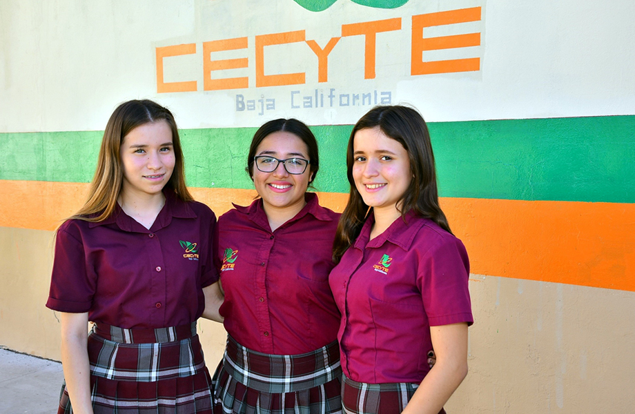 Tres estudiantes del CECyTE con uniforme guinda posando frente a una pared con el logotipo de CECyTE Baja California