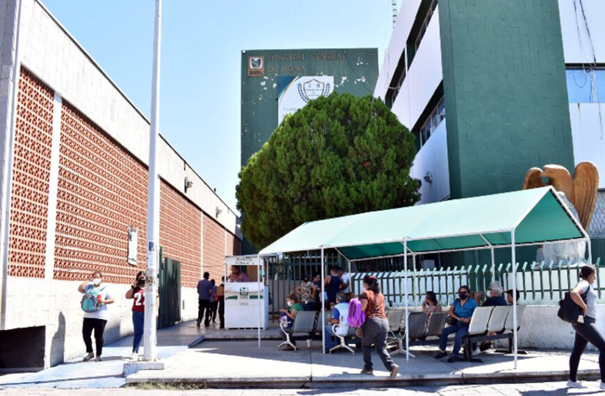 Entrada del Hospital General de Zona del IMSS en Nuevo Laredo, con personas sentadas y de pie bajo una estructura con techo verde mientras esperan ser atendidas.