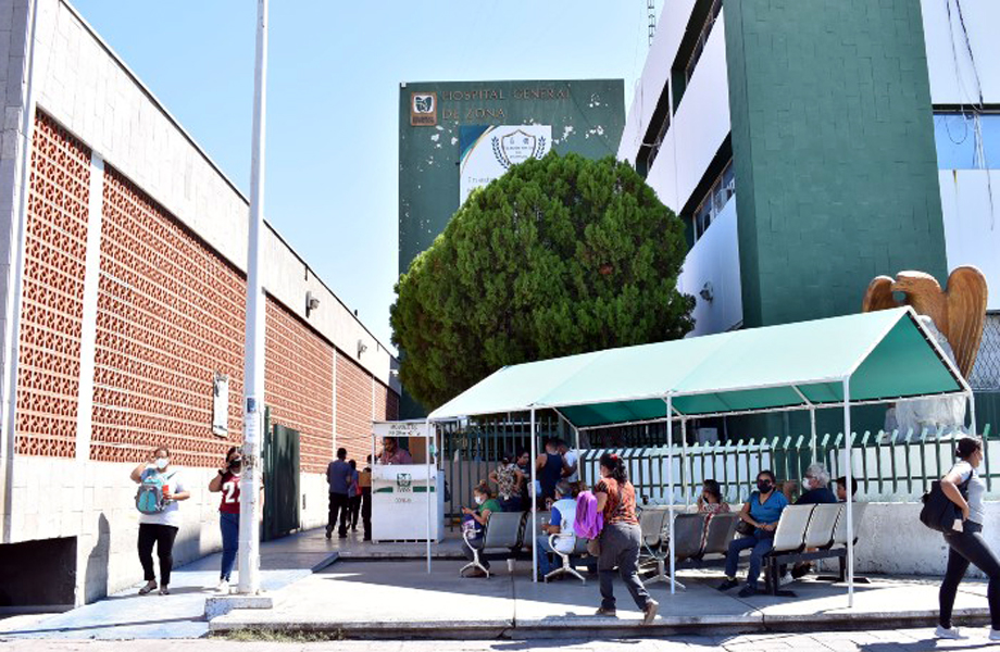 Entrada del Hospital General de Zona del IMSS en Nuevo Laredo, con personas sentadas y de pie bajo una estructura con techo verde mientras esperan ser atendidas.