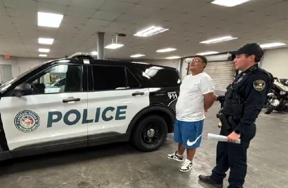 Hombre con las manos esposadas detrás de la espalda de pie junto a una patrulla de la Policía de Laredo, bajo la vigilancia de un oficial.