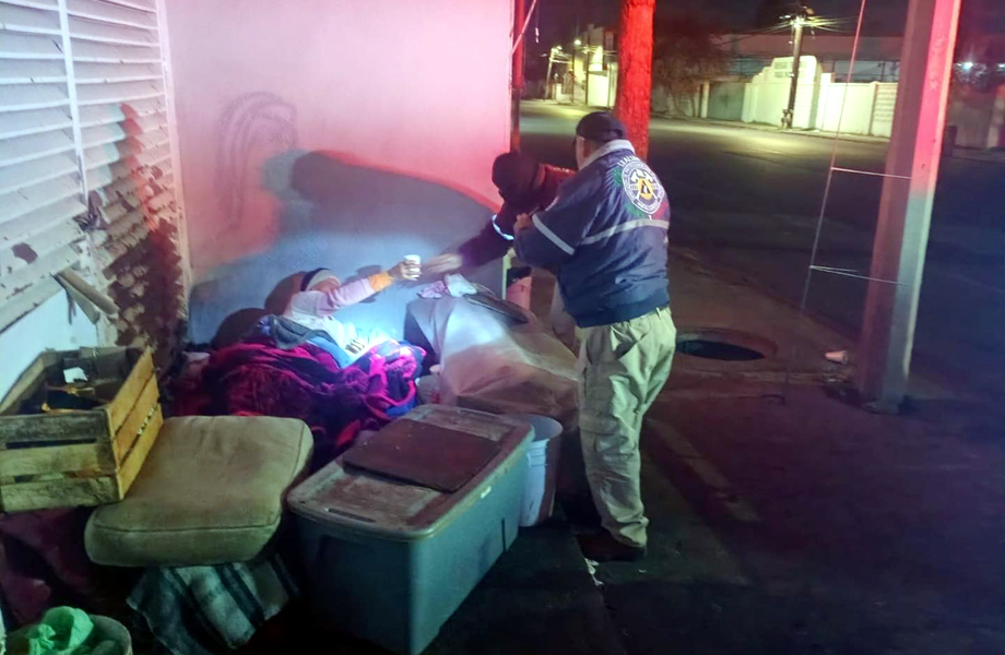 Miembro de Proteccin Civil entrega caf a una persona vulnerable que se encuentra cubierta con mantas sobre la banqueta en una calle de Nuevo Laredo durante la noche