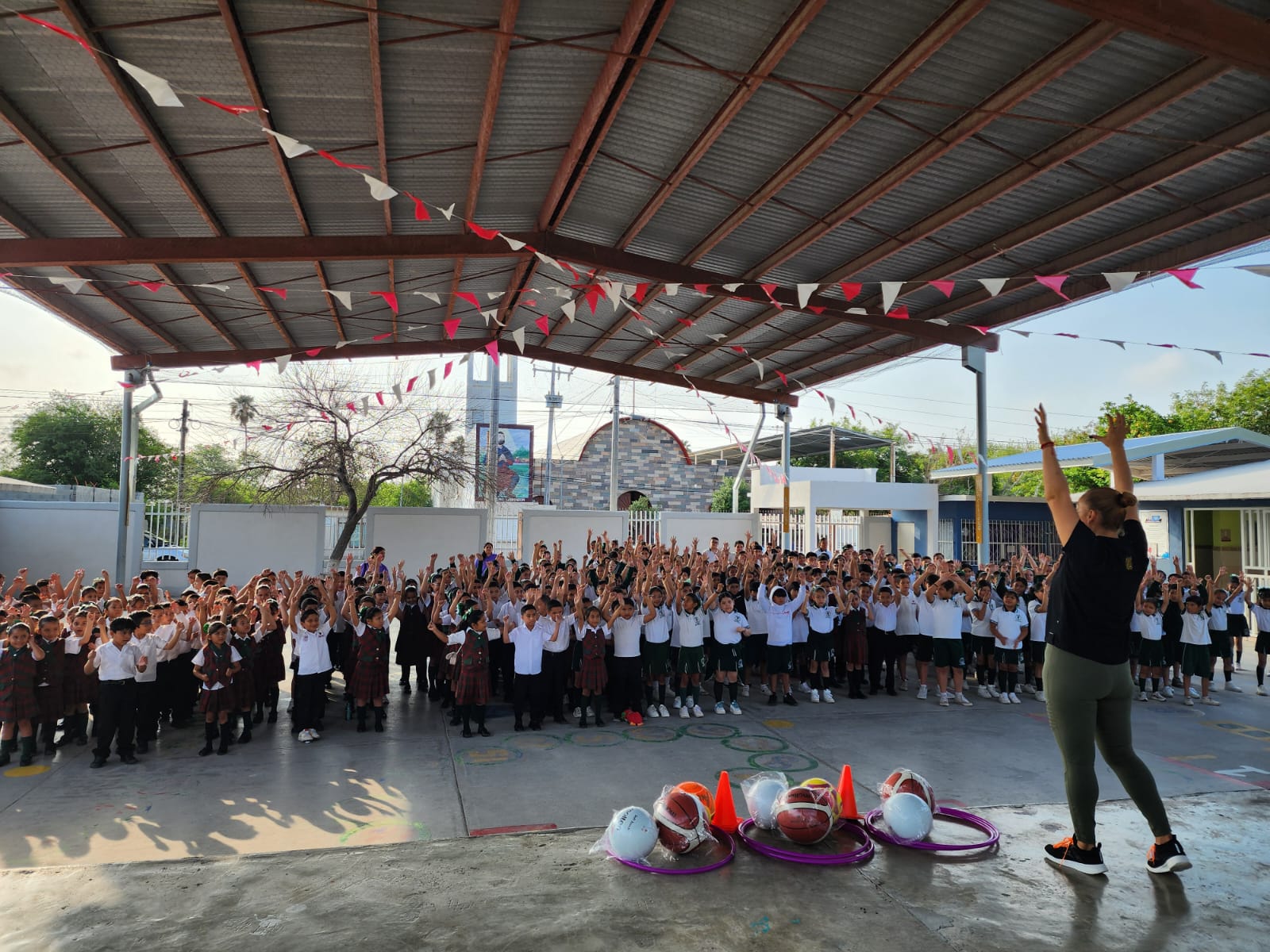 Teacher leading a large outdoor school assembly as students in uniforms raise their hands under a covered courtyard with bunting overhead: gym-like activity with balls and hoops on the ground