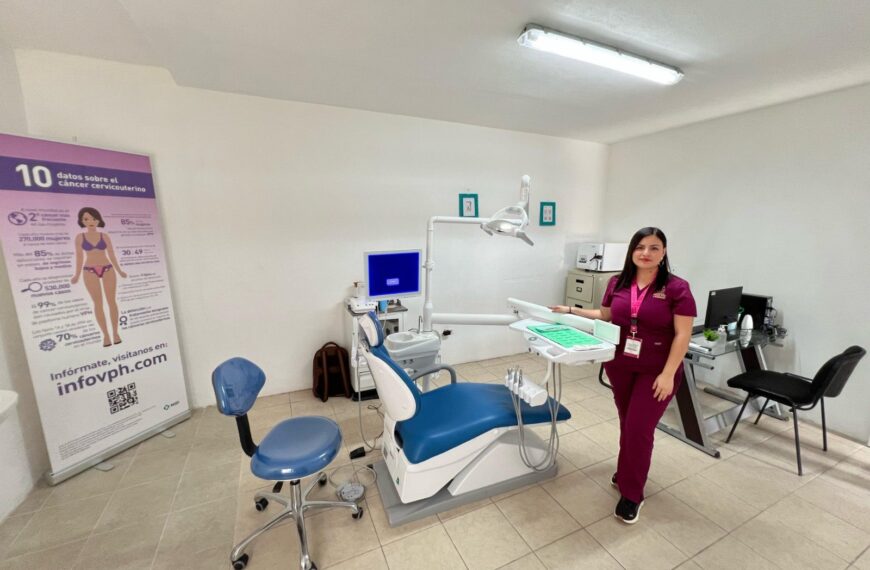 Dental treatment room with a blue and white dentist chair, overhead light, and a clinician in magenta scrubs beside a workstation.