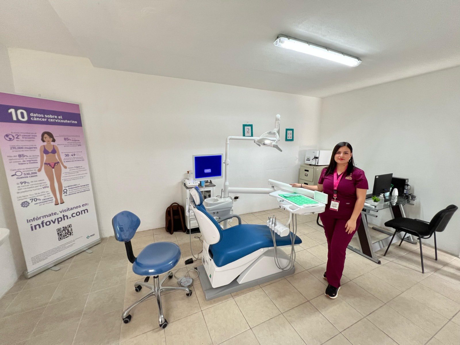 Dental treatment room with a blue and white dentist chair, overhead light, and a clinician in magenta scrubs beside a workstation.