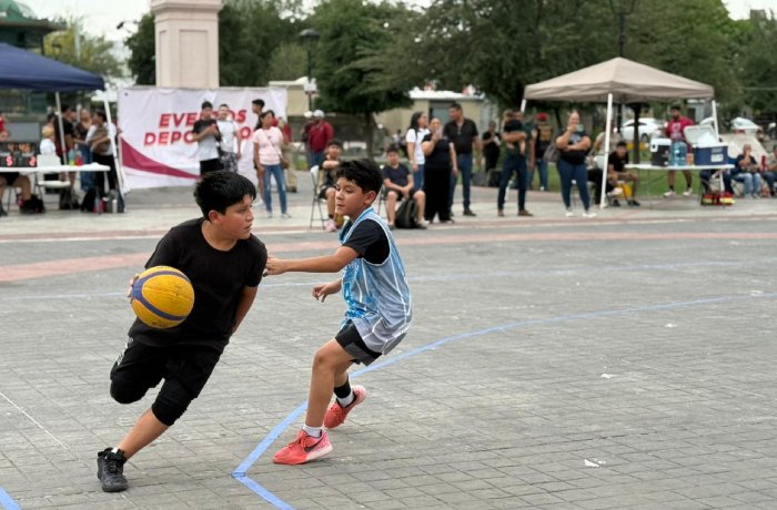 Two boys play basketball on an outdoor court at a community event; one dribbles a yellow ball while the other guards, with tents and spectators in the background.