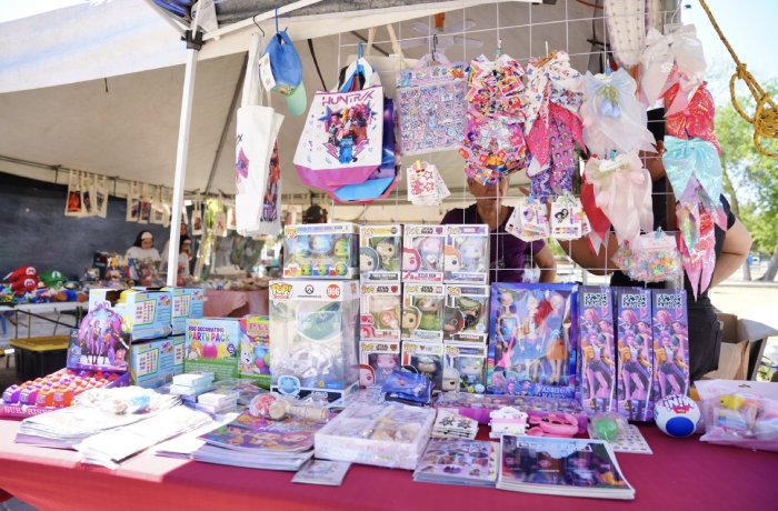 Toy stall at a market with anime figures, backpacks, and colorful bags hanging above the table.