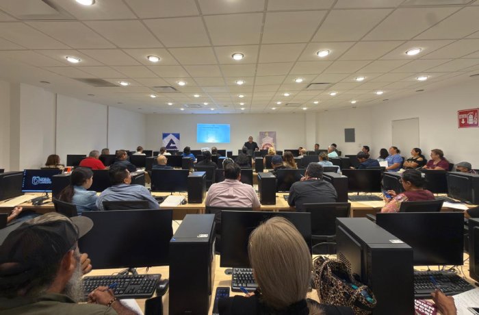 Audience seated at computer desks in a classroom-style conference room watching a presenter at the front with a projection screen above.