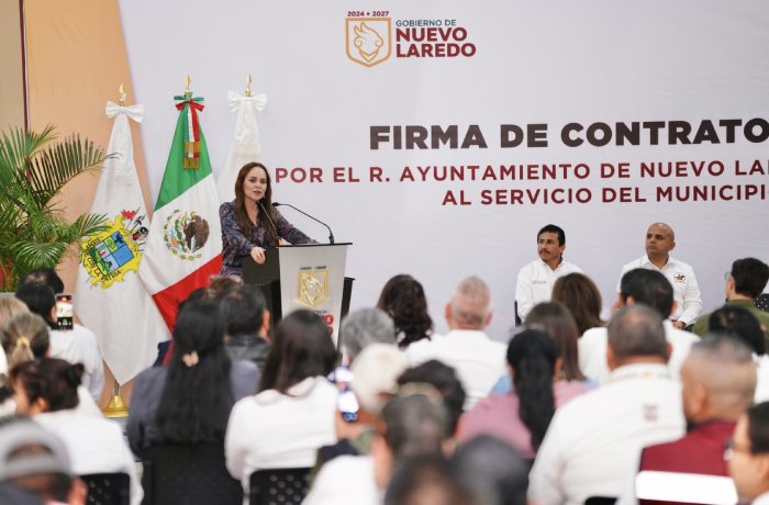 Woman at a podium addressing attendees at a contract-signing ceremony for the Nuevo Laredo city government, with flags and stage officials in the background.