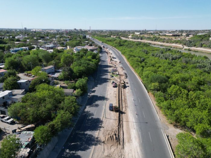 Aerial view of a highway under construction with heavy machinery and a long trench, bordered by trees and a residential area on the left