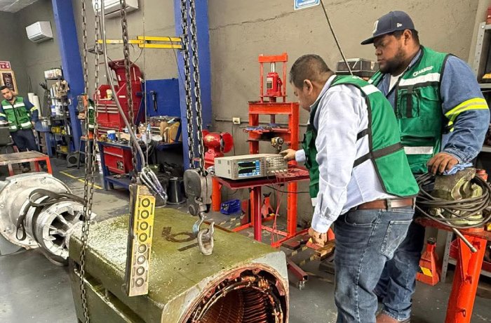 Two technicians in green safety vests inspect a large industrial motor or turbine on a workshop floor, with testing equipment and chains nearby.