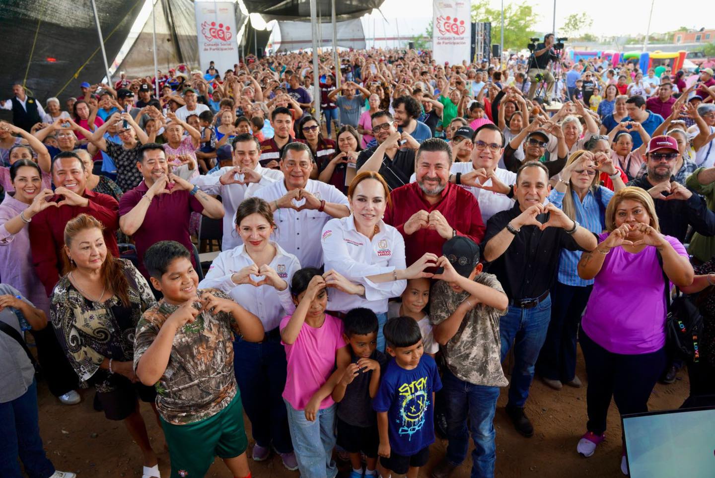 Large diverse crowd outdoors, many forming heart shapes with hands to show unity at a community event, with a few organizers in white shirts at the front smiling.