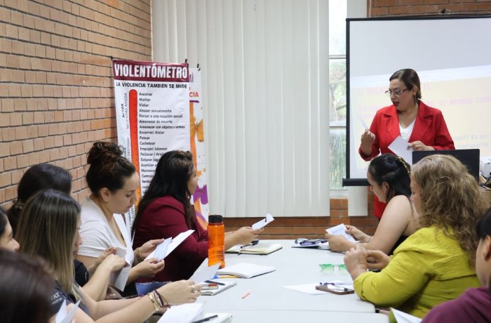 Instructor in a red blazer leads a workshop, standing near a projector as adults around a table exchange papers. A 'Violentómetro' poster is on the brick wall behind them.