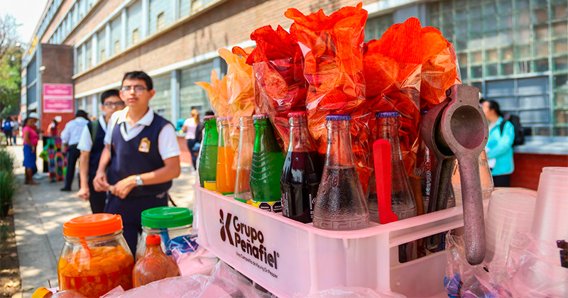 Bottled drinks on a white crate at a school fundraiser, with bright red tissue and students in uniform in the background and the crate labeled Grupo Penafiel