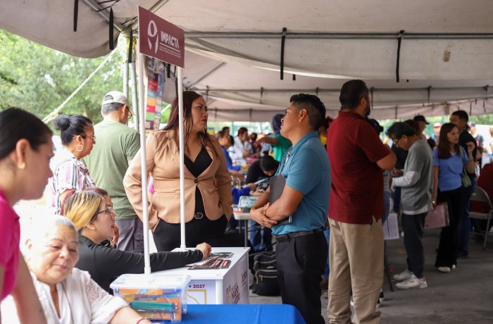 Crowded outdoor community fair under a tent with volunteers at a table welcoming attendees and sharing information, a sign reading 'Impacta' visible overhead.