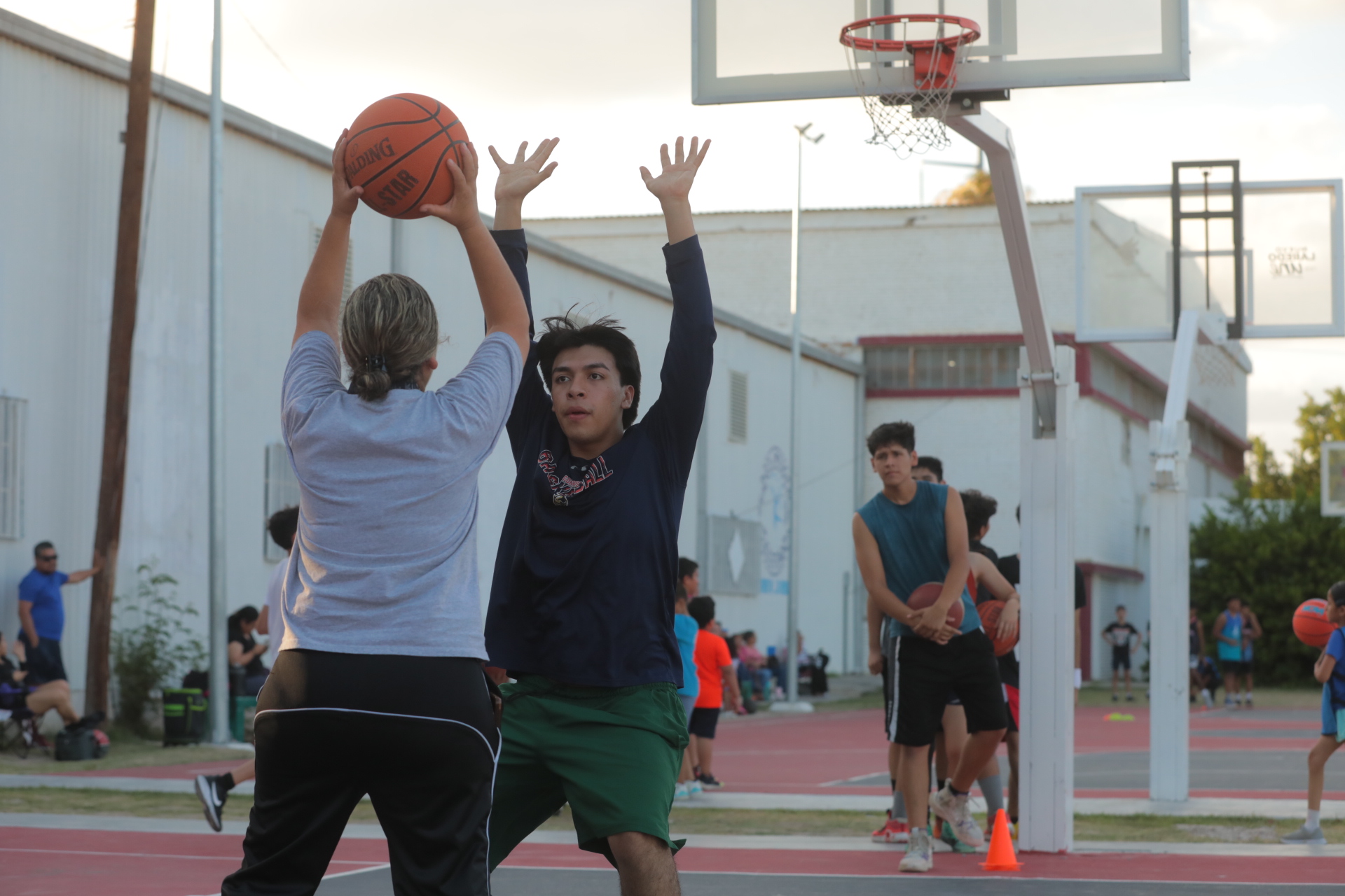 Two players jump to block a basketball shot on an outdoor court, with bystanders in the background.