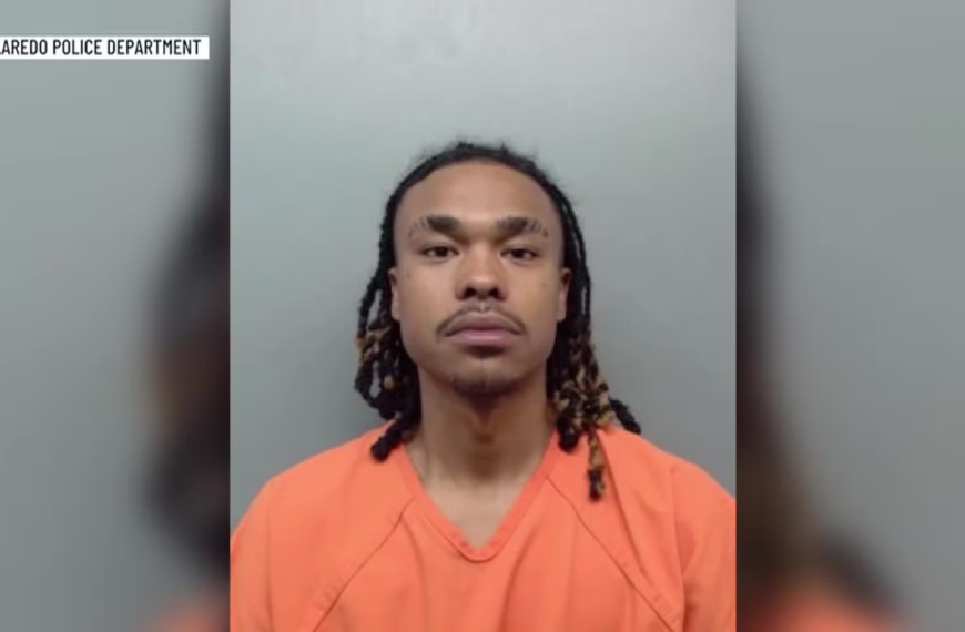Mugshot of a man with braided hair wearing an orange jail jumpsuit against a gray wall, Laredo Police Department label visible on top left.