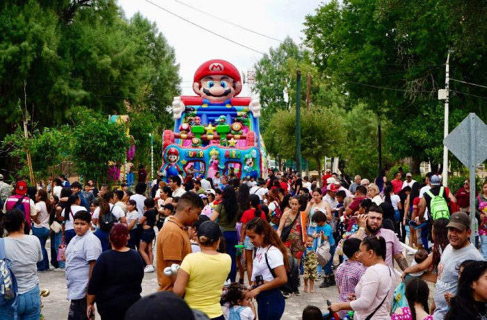 Crowded street festival with a large Mario-themed inflatable slide in the background and families watching children play nearby.