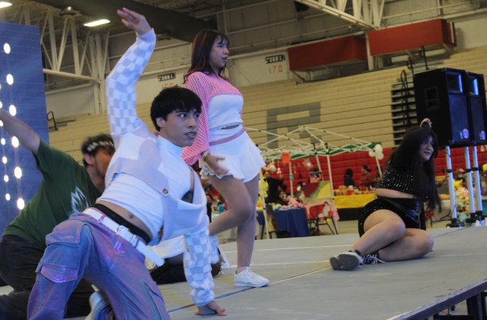 Dancers perform on a stage in a gymnasium; a man in a white jacket strikes a pose while a woman in pink and white stands beside him, and another dancer sits on the floor in black attire in the background.