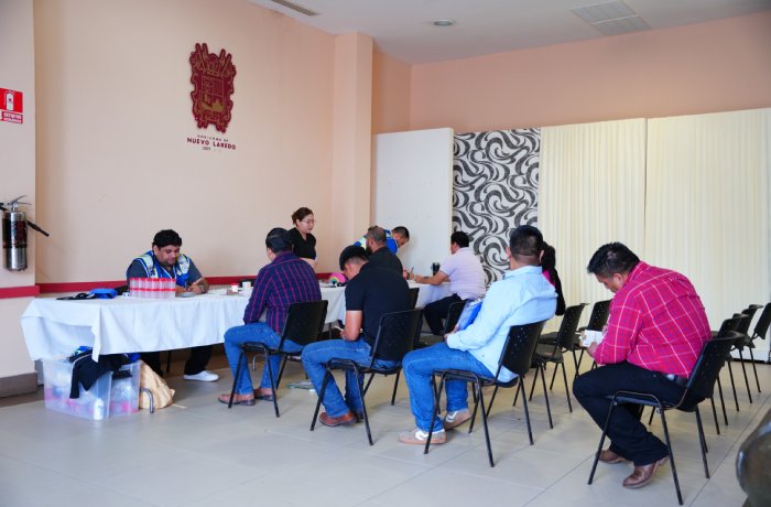 Group of people seated around a long table with a staff member assisting them with forms in a community center room with a crest on the wall