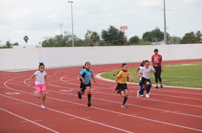 Children sprint on a red running track during practice, with a coach watching in the background.