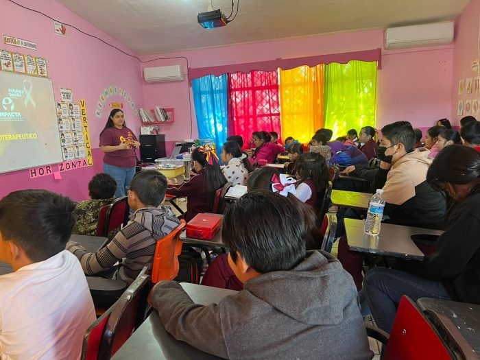 Teacher explaining a lesson at the front of a pink classroom to a crowded group of students at desks; rainbow curtains light the window.