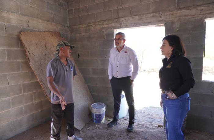 Three adults stand inside a partially built cinder-block room, conversing; a worker in a cap explains something while a man in a white shirt and a woman in jeans listen, with a bucket and plywood panel nearby.