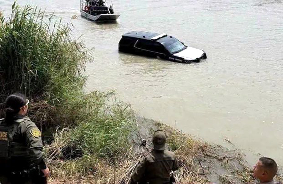 Police and responders stand on riverbank watching a partially submerged black car in a murky river.