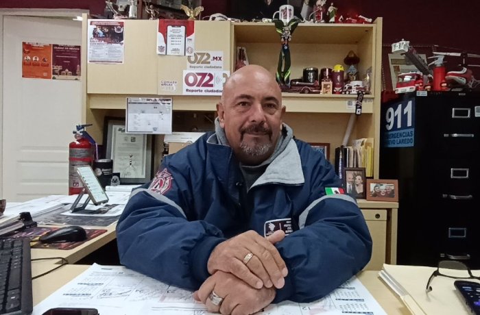 Bald man with a goatee sitting at a desk in a cluttered office, wearing a blue jacket with patches.