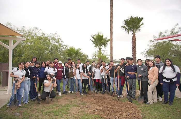 Group of people posing with shovels after a tree-planting event outdoors, with palm trees in the background and a dug dirt patch in front.