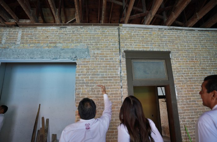 Three people in white shirts stand indoors at a construction site, pointing at a brick wall beneath exposed ceiling beams and a doorway to the right.