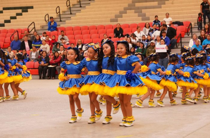 Young girls in blue and yellow costumes perform a synchronized dance on a gymnasium floor as an audience watches.