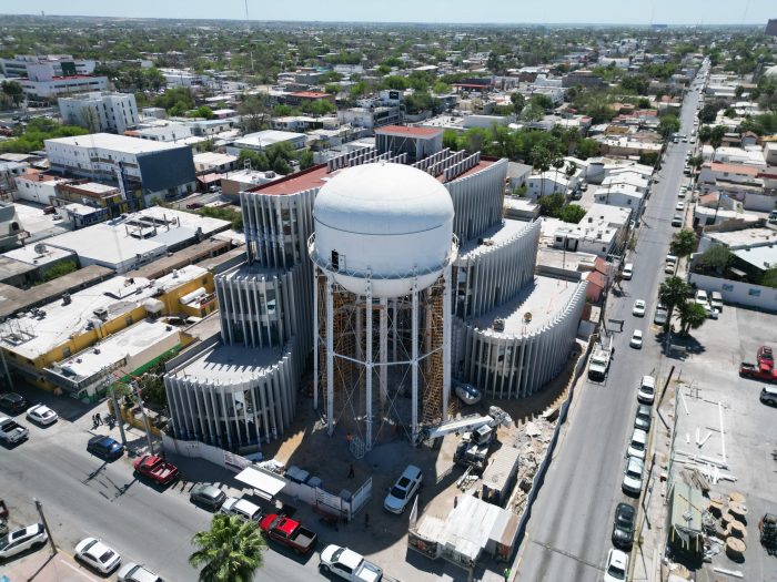 Aerial view of a large white water storage tank on a circular elevated structure under construction in a residential city block.