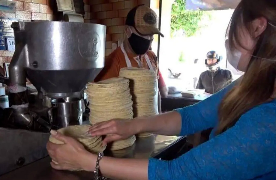 Workers in a tortilla shop wearing masks stack freshly made tortillas by a metal press.