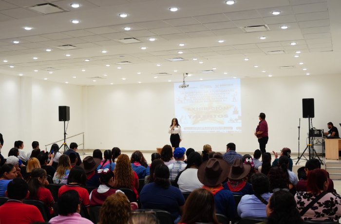 Woman speaking on a stage to a large seated audience in a bright conference room, with a projection screen behind her.