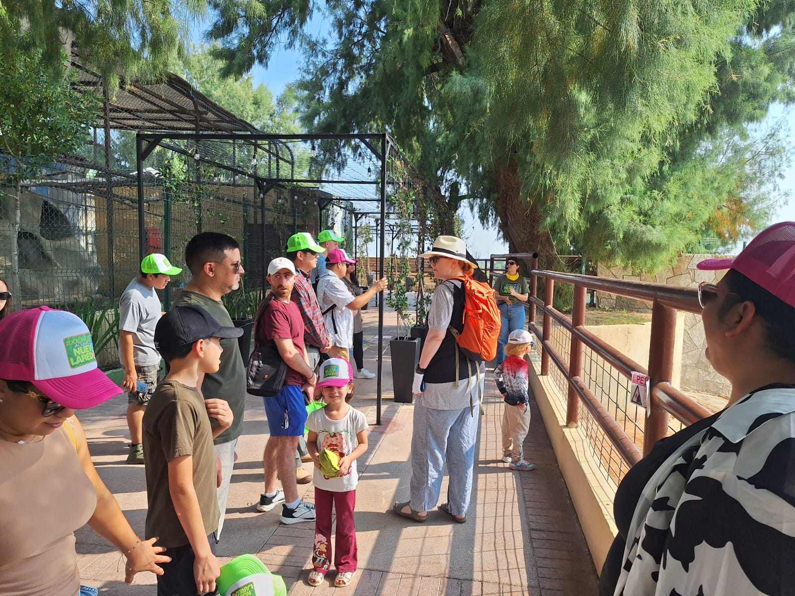 A group of people, including children, stand in a shaded outdoor area along a railing, wearing hats and backpacks on a sunny day at a park or zoo.