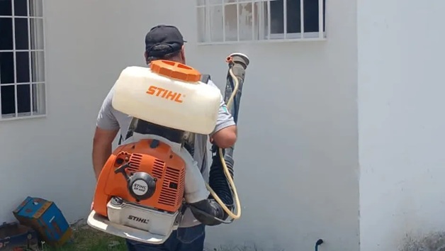 Worker wearing a STIHL backpack sprayer and holding the hose, ready to spray near a white wall.