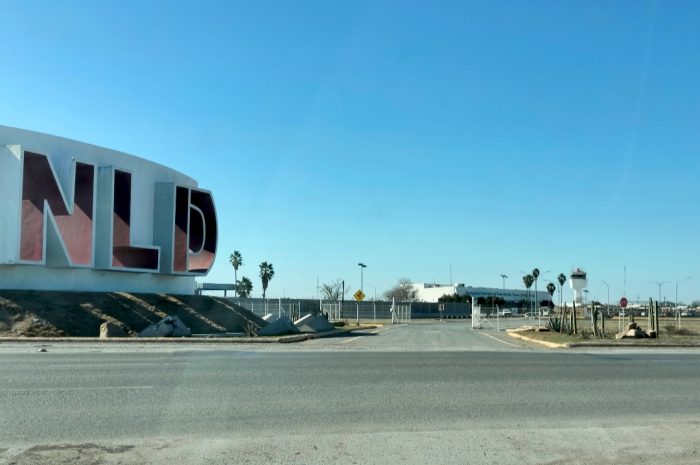 Wide street view with a large curved sign reading 'NLD' on the left, palm trees, and a gated facility in the distance.