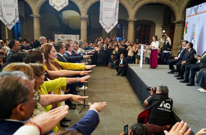 Dignitaries and audience extend their arms in a ceremonial pledge in a grand arched hall, with a speaker at the podium and photographers nearby.
