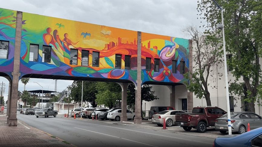 Colorful mural on the side of a two-story building with abstract swirls and a city skyline, beside a street with parked cars and trees.