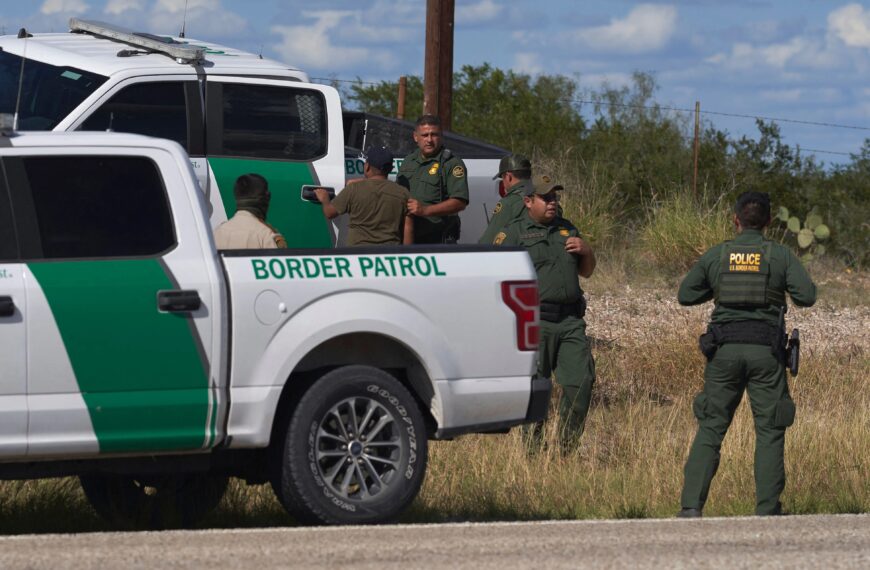 Border Patrol SUV with green stripe and agents standing in a grassy field near a dirt road.